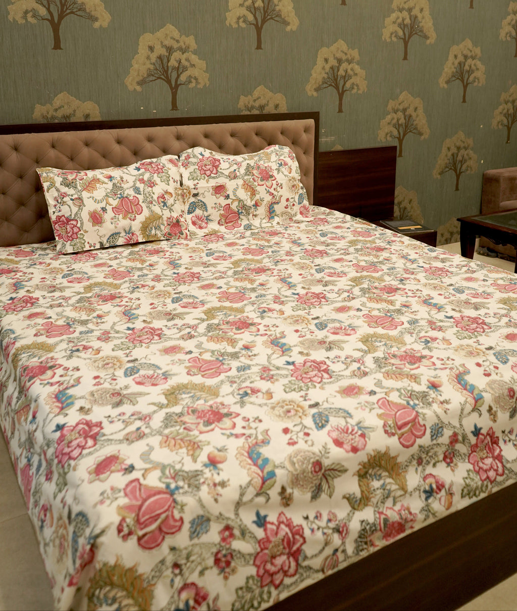 A bed dressed in a floral quilt and matching pillow shams, set against a wall with matching tree-patterned wallpaper. The bed frame is dark wood, and a small bedside table is visible on the right.