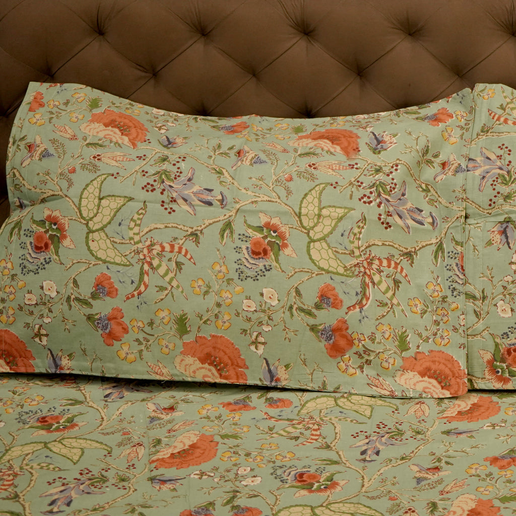 A close-up of a bedding set featuring a sage green floral and botanical print with orange, red, and purple flowers, styled on a bed with a brown tufted headboard.