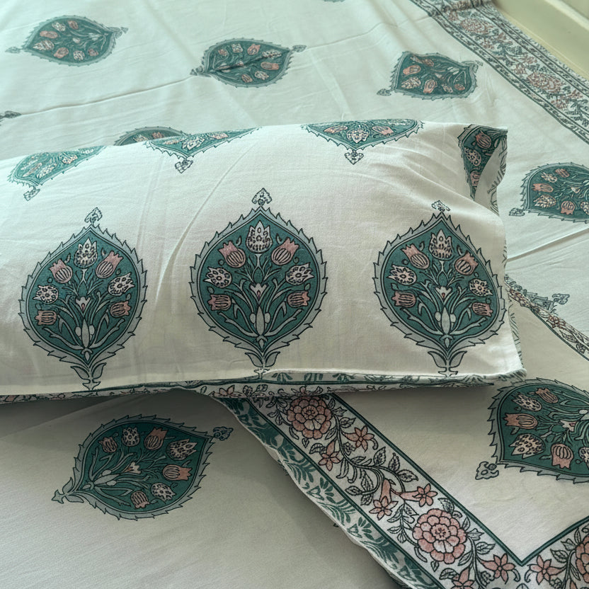 A bed made with a white cotton block-print bedding set featuring teal and pink floral motifs, with two matching pillowcases and a decorative book placed on top.