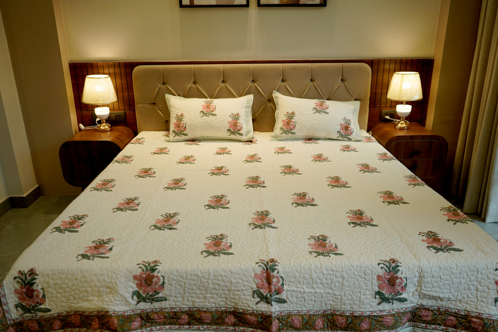 A neatly made bed in a bedroom setting, featuring a quilted cream-colored bedspread and matching pillowcases with a repeating pink rose and green leaf pattern. The bed is flanked by two dark wood nightstands with lit table lamps, set against a tufted beige headboard.